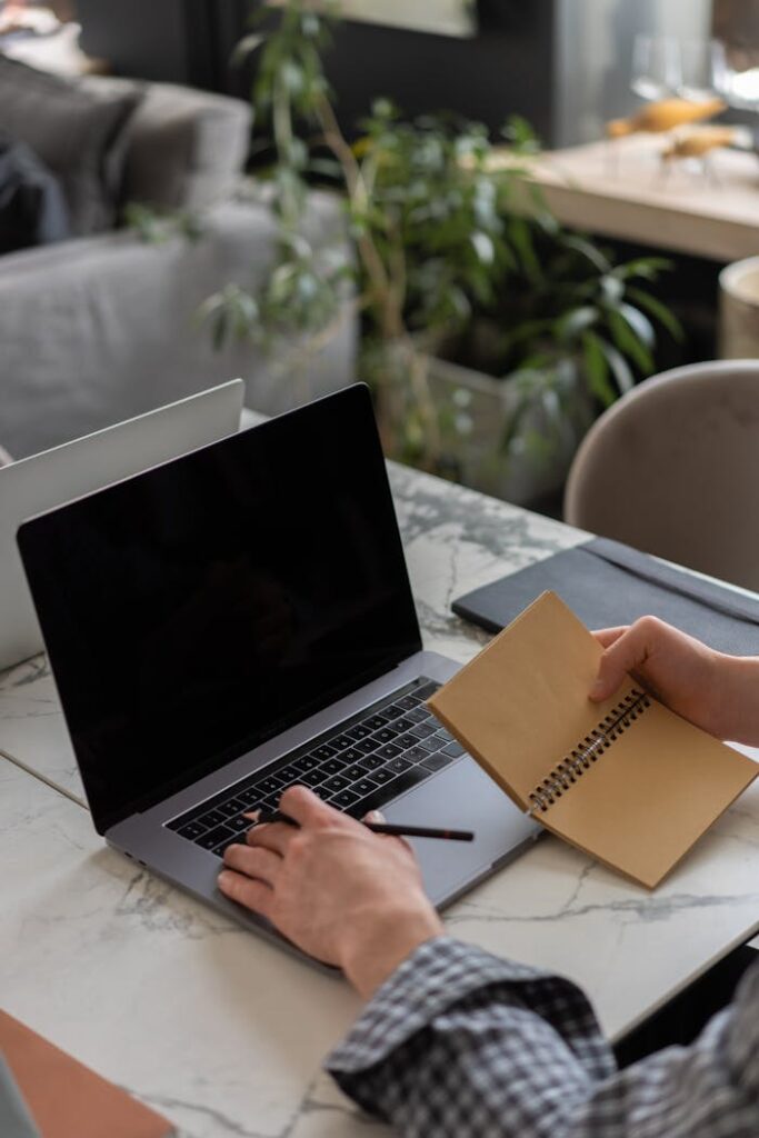 Close-up of hands writing in a notebook while using a laptop in a cozy indoor office setting.